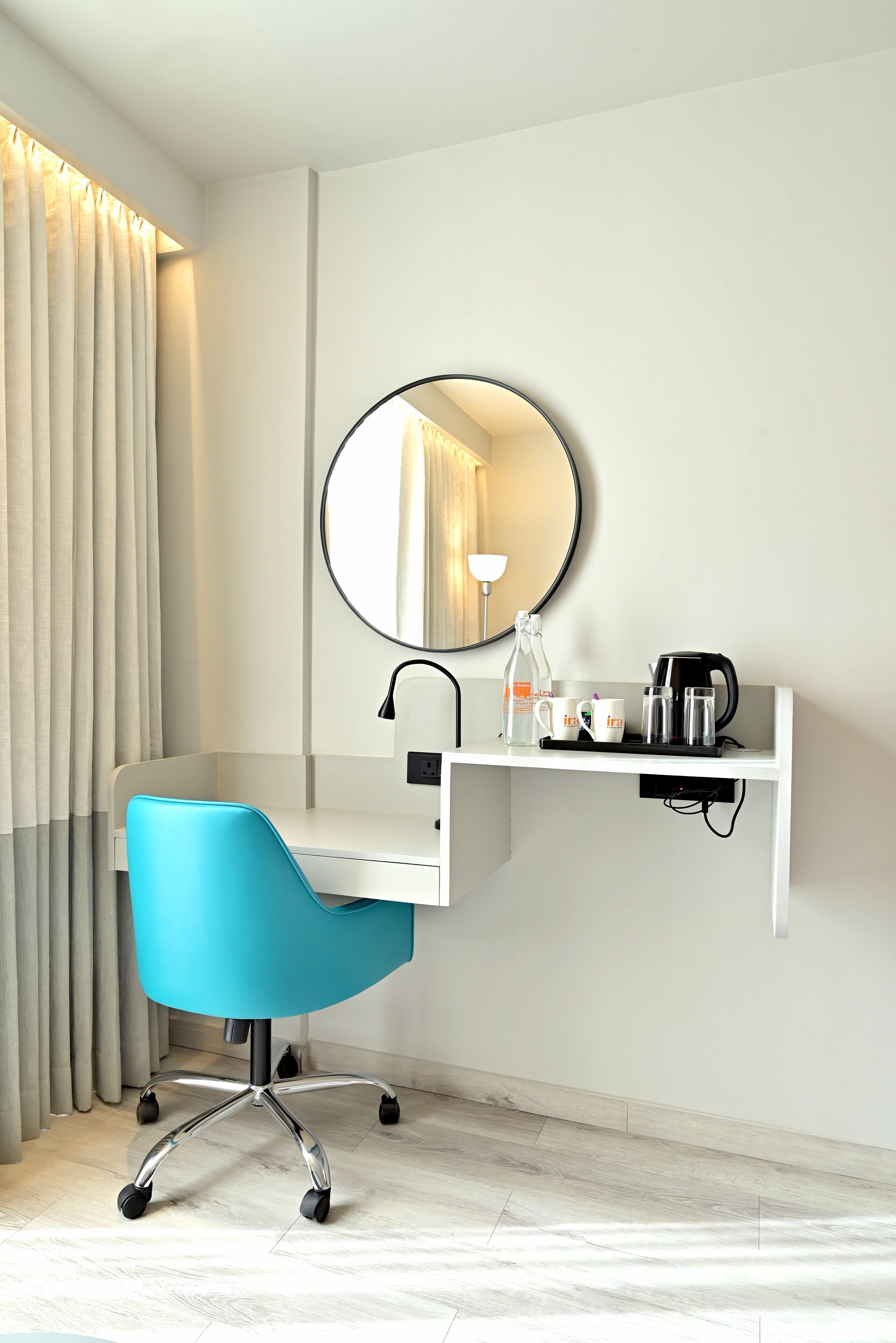 A modern hotel room with a white desk, a blue office chair, a round mirror, a coffee maker, and a bottle of water.