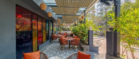 An outdoor dining area with wicker chairs and tables, a glass ceiling, and potted plants.