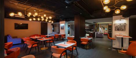 Un intérieur moderne avec des chaises et des tables orange, un plafond orné de luminaires suspendus et des œuvres d’art accrochées aux murs.