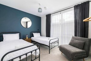 Hotellihuone, jossa on kaksi sänkyä, pyöreä peili, yöpöytä lampun kanssa, harmaa sohva ja suuri ikkuna, jossa on verhot.