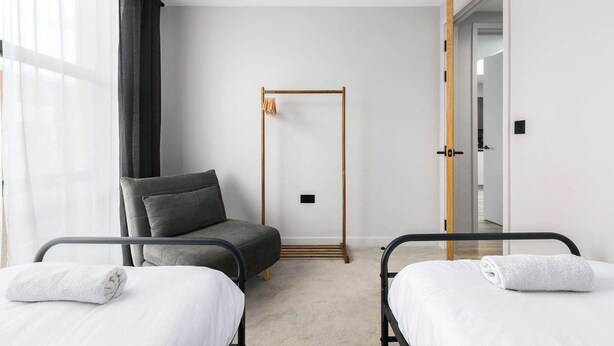Ein Hotelzimmer mit zwei Betten, einem Sessel, einem Holzschrank und einer Tür zu einem anderen Raum.