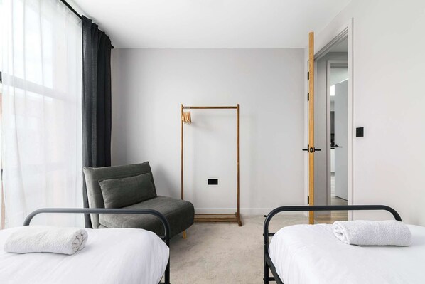 A hotel room with two beds, a chair, a wooden wardrobe, and a door leading to another room.