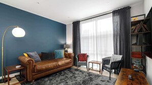 Ein Wohnzimmer mit braunem Ledersofa, einem kleinen Beistelltisch, einer Lampe, einem Bücherregal und einem großen Fenster mit Vorhängen.