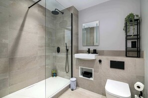Moderni kylpyhuone, jossa on lasiseinäinen suihku, valkoinen pesuallas, wc ja pieni hylly, jossa on hygieniatuotteita.