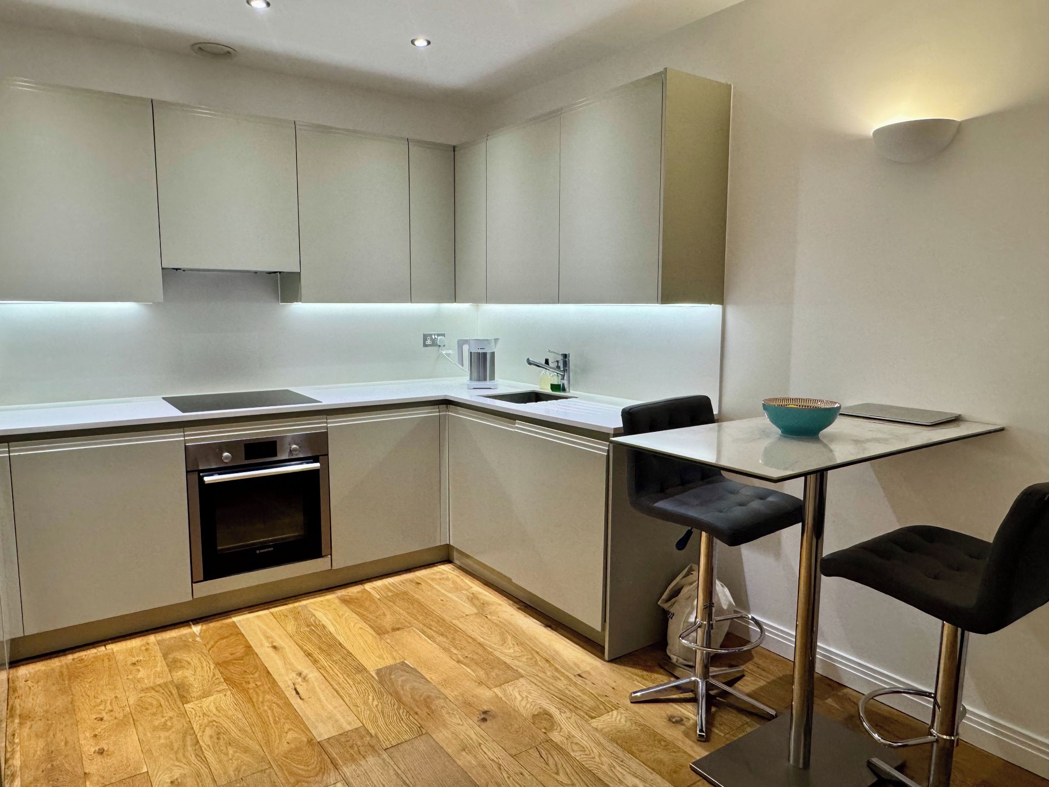 A modern kitchen with a built-in oven, a sink, and a coffee maker. There is a small table with two chairs and a bowl on it.