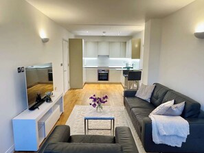 A modern living room with a flat-screen TV, a sofa, a coffee table, and a kitchen in the background. - Designer 1 Bed Flat in Iconic Building (Perivale)