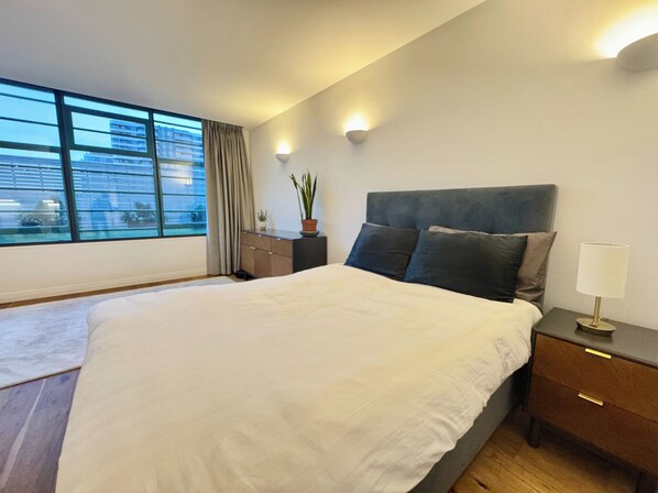 A modern bedroom with a large bed, a nightstand, a lamp, a potted plant, and a view of buildings through the window.
