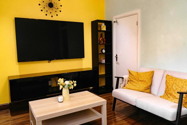 Un salón con un televisor de pantalla plana montado en la pared, una mesa de centro de madera con un jarrón de flores, un sofá blanco con cojines amarillos y un mueble de entretenimiento negro con estantes.
