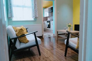 Una sala moderna con sofá y sillón, una pequeña mesa de comedor, y una cocina al fondo.