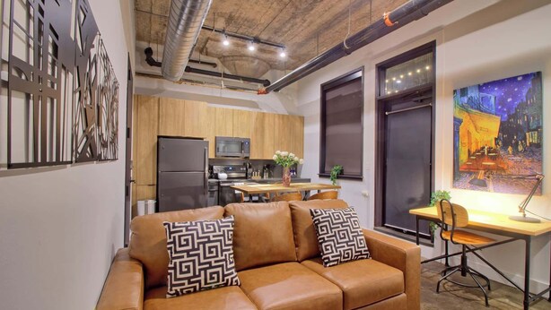 A modern living room with a brown leather sofa, a wooden dining table, and a kitchen in the background. - Chic Ultramodern Loft Suite w/ Designer Features (St. Louis)