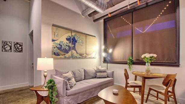 A modern living room with a sofa, coffee table, and chairs, a large window with curtains, and a wall art piece. - Chic Ultramodern Minimalist Loft Suite + Patio (Saint Louis)