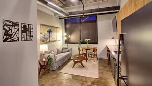 A modern living room with a sofa, coffee table, and dining table. There is a large window with a view, a ceiling fan, and a refrigerator. - Chic Ultramodern Minimalist Loft Suite + Patio (Saint Louis)