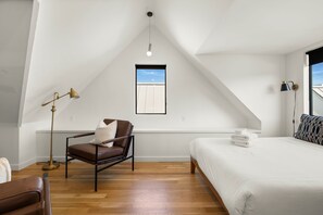 Un dormitorio con techo inclinado, una ventana, cama con ropa de cama blanca, una silla de cuero marrón y una lámpara de pie.