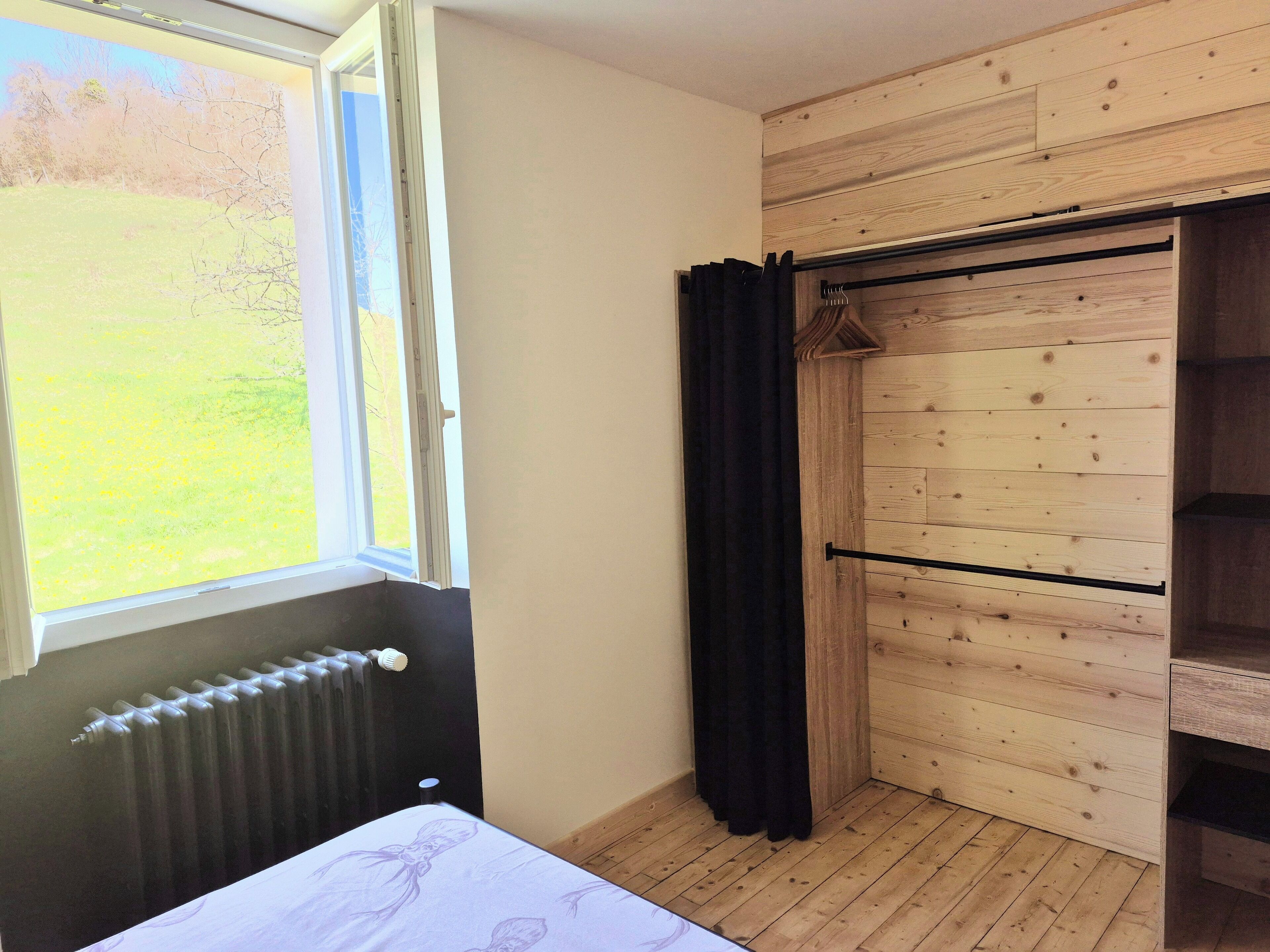 Ein Zimmer mit einem Bett, einem Heizkörper, einem Fenster mit Blick auf ein Wiesenfeld, einem Holzschrank und einem schwarzen Vorhang.