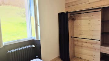 Ein Zimmer mit einem Bett, einem Heizkörper, einem Fenster mit Blick auf ein Wiesenfeld, einem Holzschrank und einem schwarzen Vorhang.