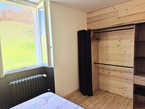 Une pièce comprenant un lit, un radiateur, une fenêtre donnant sur un champ verdoyant, une armoire en bois et un rideau noir.