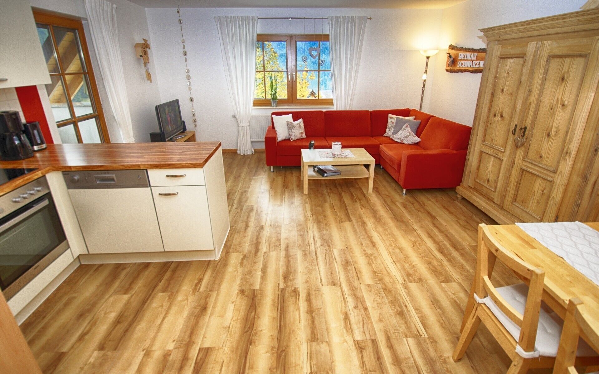 Un ambiente compatto con angolo cottura, un divano rosso, un tavolo da pranzo in legno e una finestra con tende.