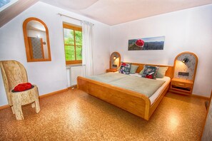Un dormitorio con una cama de madera, una silla, un espejo, una ventana y un cuadro colgado en la pared.