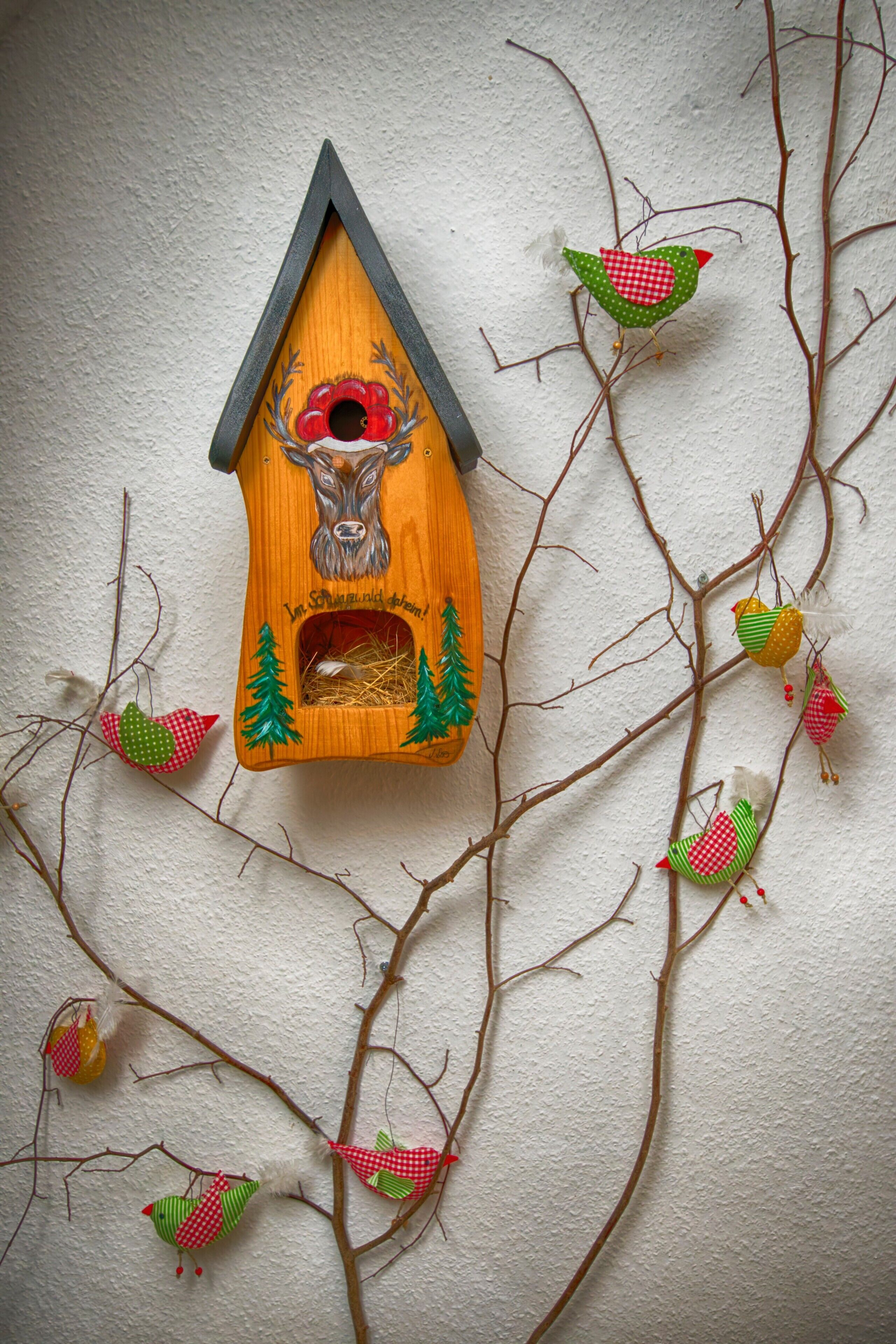Ein Vogelhaus aus Holz mit Hirschmotiv, umgeben von Ästen und kleinen Vögeln.