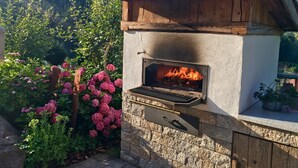 Una parrilla de piedra construida al aire libre con techo de madera, rodeada de exuberante follaje y flores rosadas.