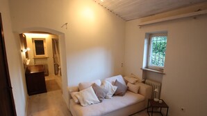 A room with a beige sofa, a small table, a radiator, and a window with a view of greenery.