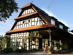 Il s’agit d’un bâtiment traditionnel à colombages, doté d’un toit en pente, d’une cheminée bien visible et d’un petit porche avec des chaises.