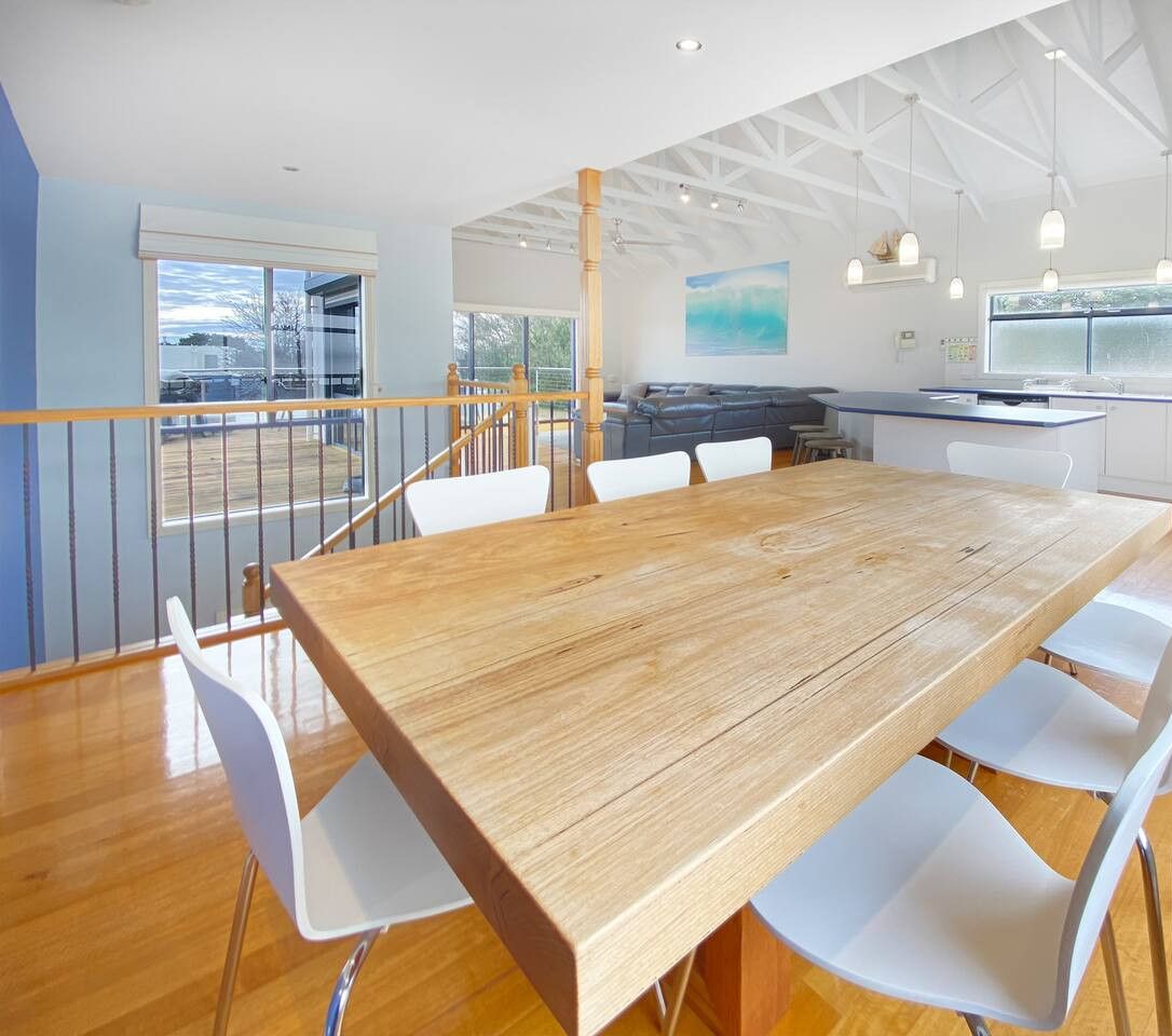 Un vaste espace de salle à manger avec une table en bois et des chaises blanches, une cuisine avec des meubles blancs et un balcon doté d’une balustrade.