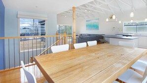Un vaste espace de salle à manger avec une table en bois et des chaises blanches, une cuisine avec des meubles blancs et un balcon doté d’une balustrade.