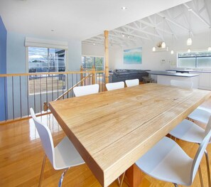Un vaste espace de salle à manger avec une table en bois et des chaises blanches, une cuisine avec des meubles blancs et un balcon doté d’une balustrade.