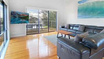 A spacious living room with a large black leather sofa, a wooden coffee table, and a painting of a wave on the wall.