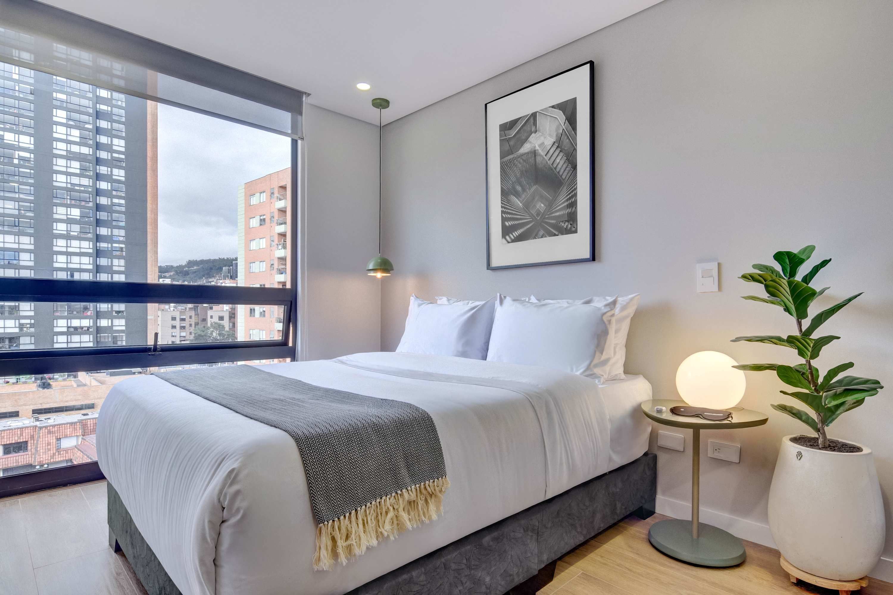 Une chambre moderne avec un grand lit, une vue sur la ville, une œuvre d’art encadrée, une lampe de chevet et une plante en pot.