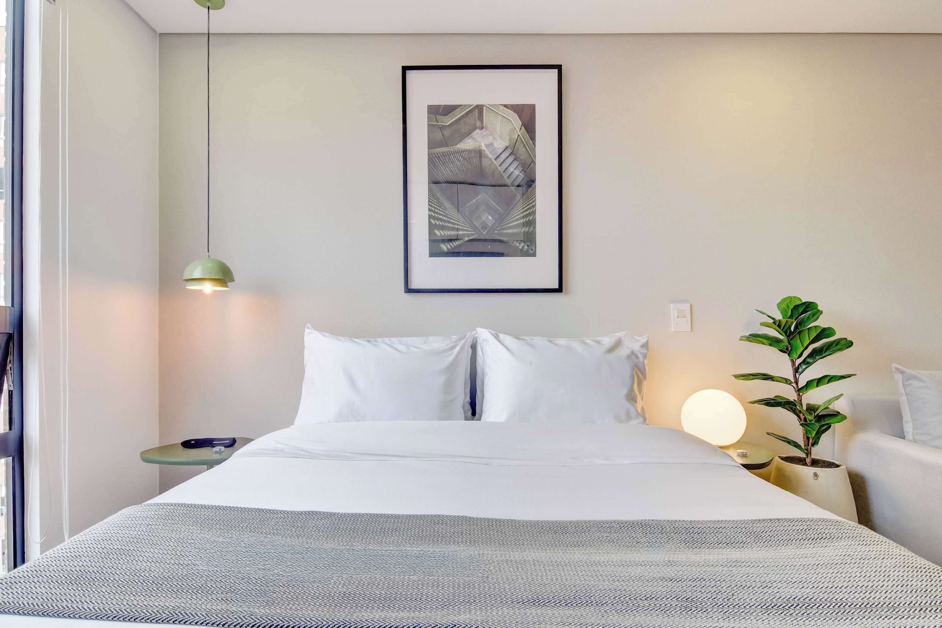 Een netjes opgemaakt bed met witte lakens, een lijst met een kunstwerk erboven, een bedlampje en een klein tafeltje met een plantje.