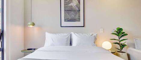 Een netjes opgemaakt bed met witte lakens, een lijst met een kunstwerk erboven, een bedlampje en een klein tafeltje met een plantje.