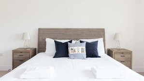 A neatly made bed with white linens and a variety of pillows, including a striped one with a building design. There are two bedside tables with lamps and a wooden headboard. - Modern Family Retreat Nr Beaches and Marina (Chichester)