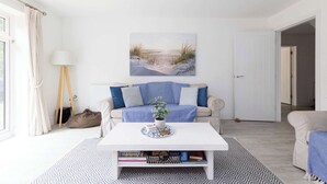 A living room with a white sofa, a coffee table, and a bookshelf. There is a painting on the wall and a lamp on a side table. - Modern Family Retreat Nr Beaches and Marina (Chichester)