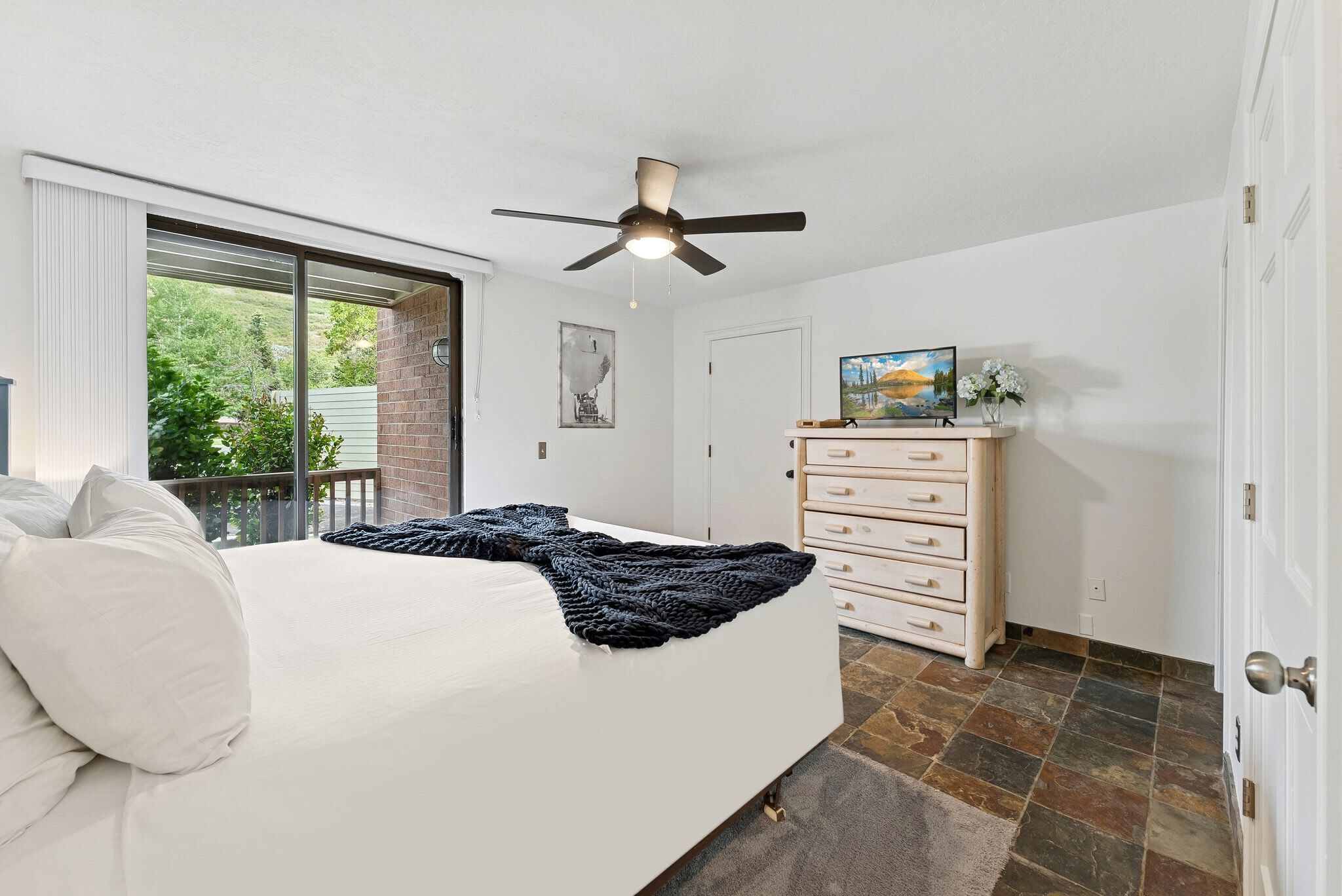 Ein Schlafzimmer mit Bett, Deckenventilator, Kommode und Fenster mit Blick ins Grüne.