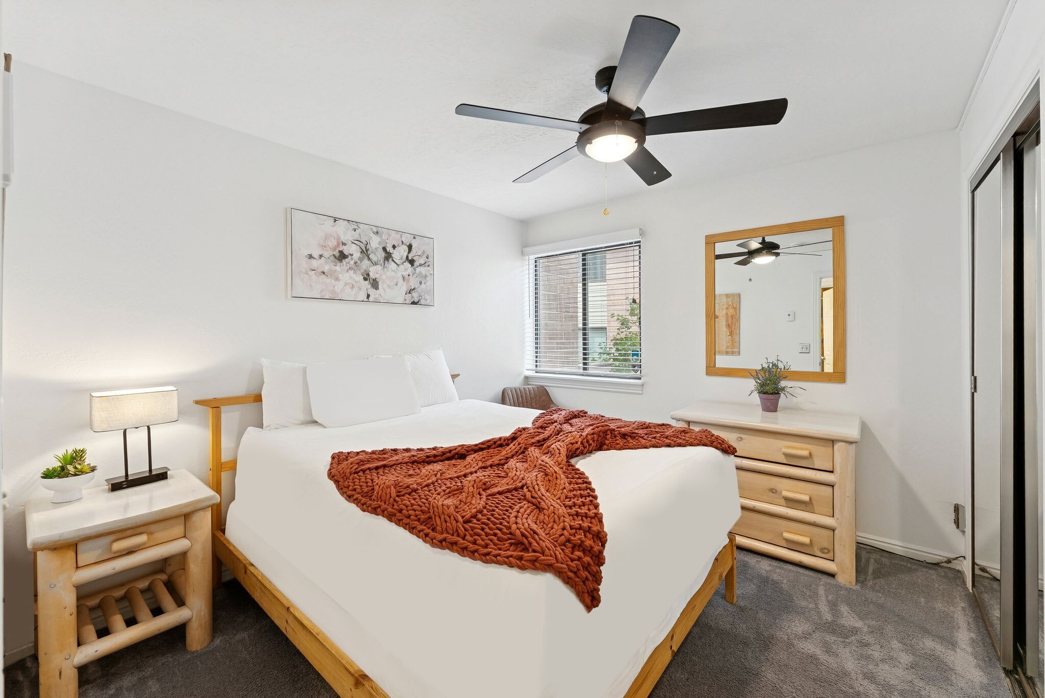Une chambre à coucher comprenant un lit, une table de chevet, une lampe, un ventilateur de plafond, une fenêtre avec stores et un miroir.