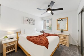 Ein Schlafzimmer mit Bett, Nachttisch, Lampe, Deckenventilator, Fenster mit Jalousien und einem Spiegel.