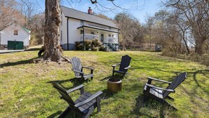 Una casa con un porche, un árbol grande y cuatro sillas Adirondack dispuestas alrededor de un brasero.