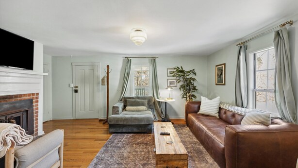 Una sala de estar con chimenea, un sofá de cuero marrón, una mesa de centro de madera, un sillón gris y un ventanal con cortinas.