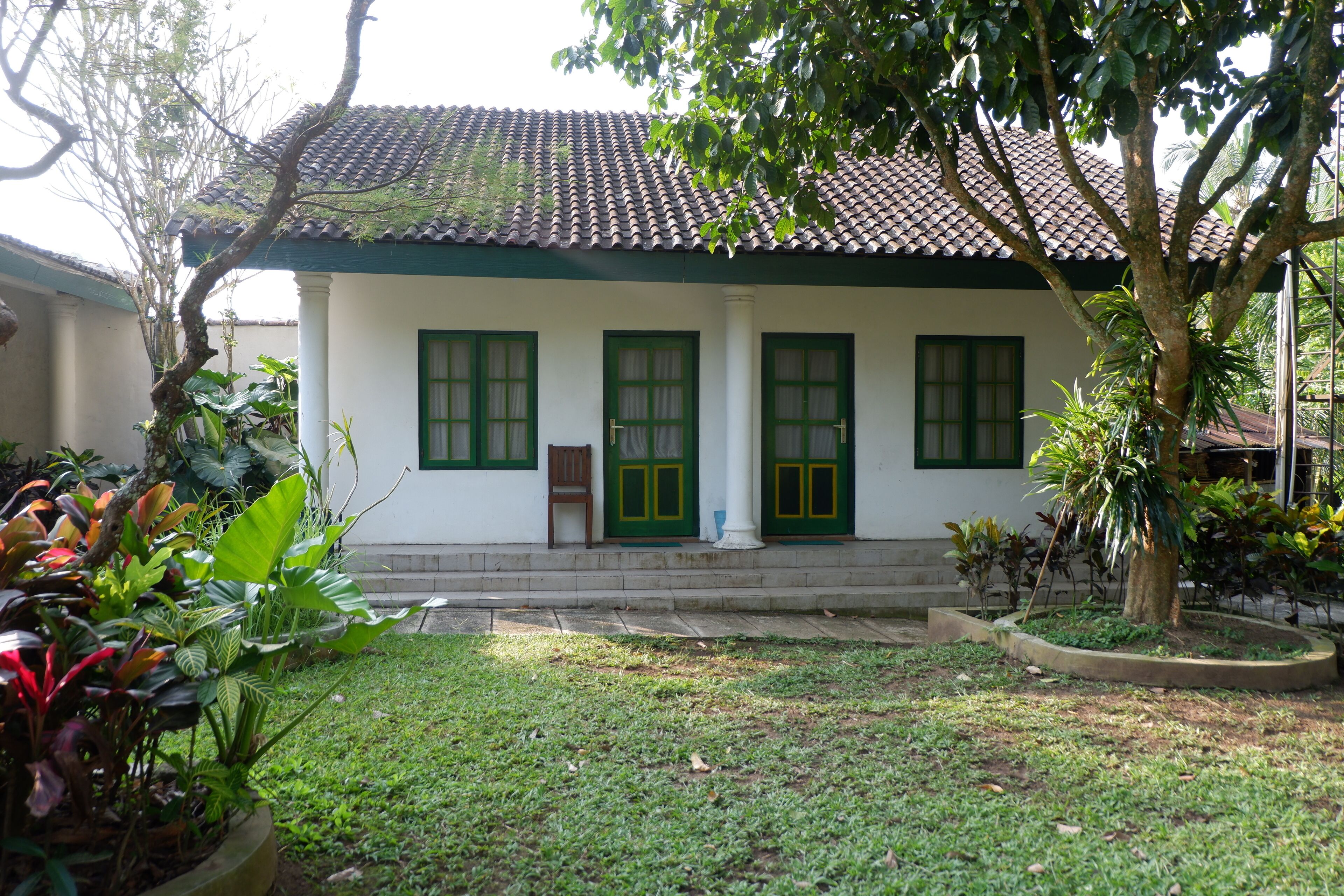 A white building with green doors and windows, surrounded by a well-maintained garden with various plants.