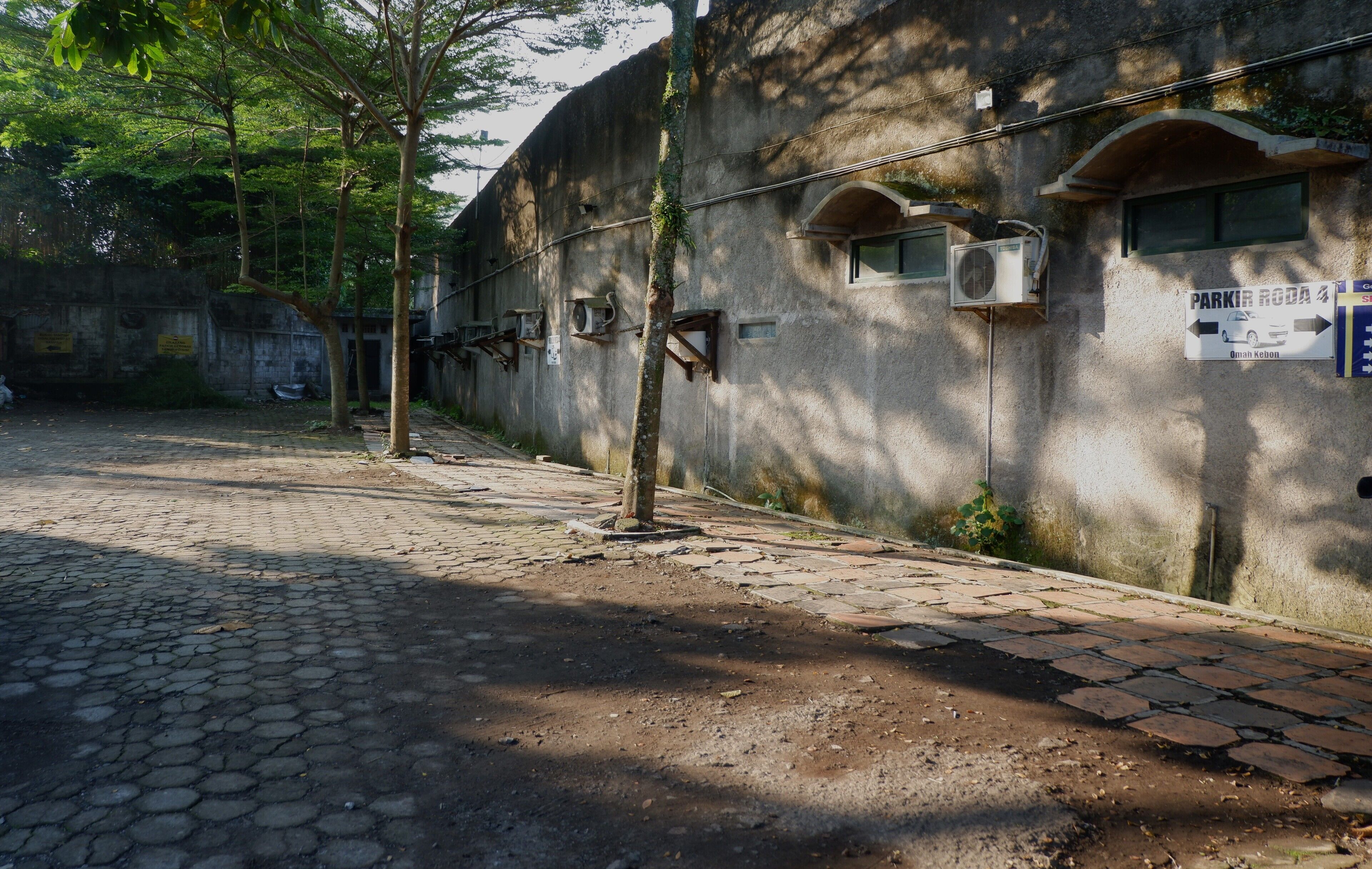A paved courtyard with a cobblestone path, a tree, and a building with air conditioning units and a sign indicating a parking area.