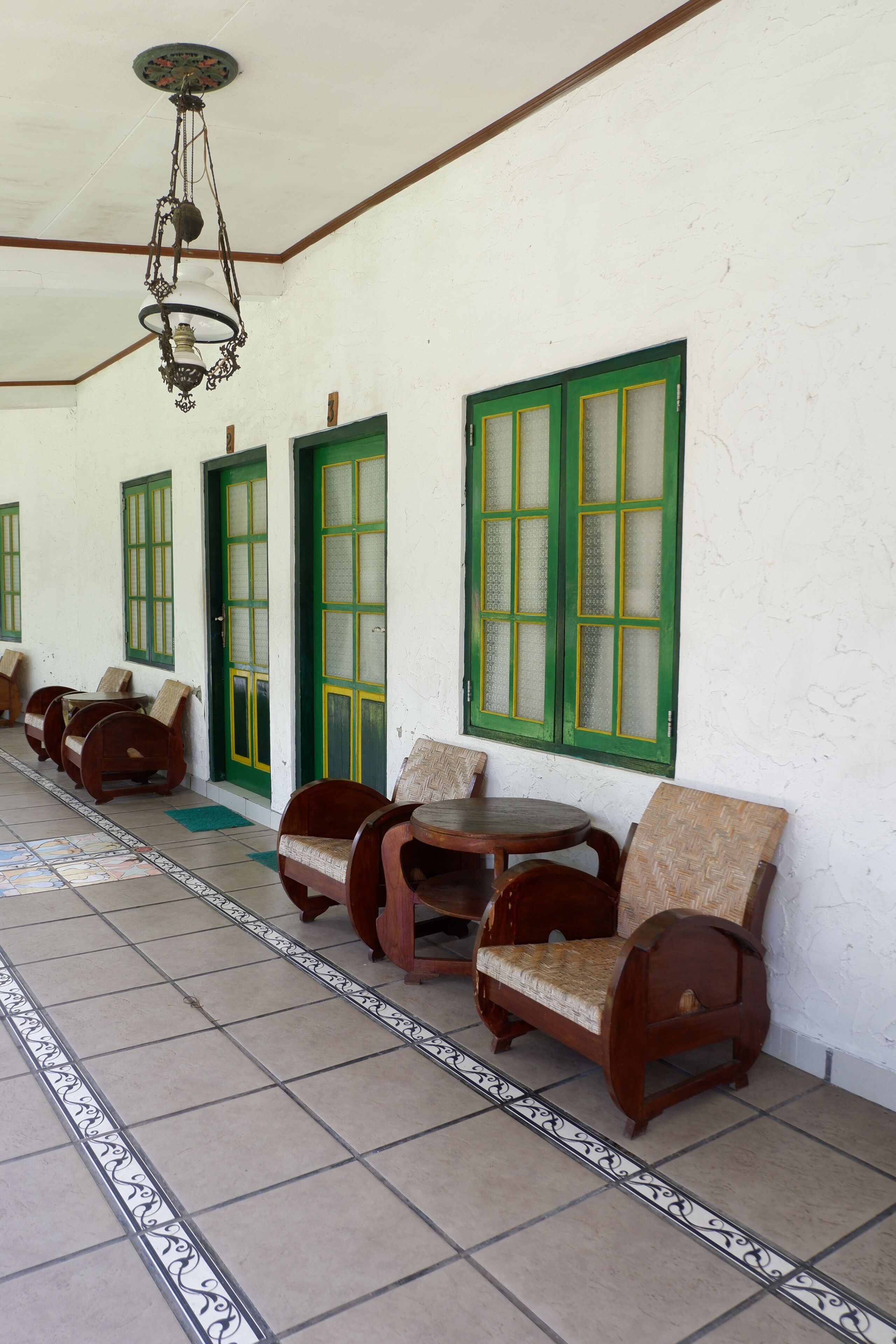 A corridor with wooden benches and chairs, green-framed doors, and a tiled floor with decorative borders.