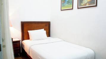 Una cama pequeña y bien hecha con cabecera de madera en una habitación con paredes blancas. Hay dos cuadros en la pared, uno que representa un paisaje y el otro, una naranja.