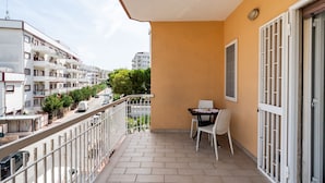 A balcony with a table and chairs, a view of a street with parked cars, and a building with multiple balconies in the background. - Roomy Family Apartment by Wonderful Italy (Bari)