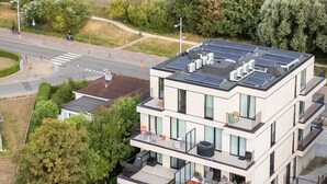 Un edificio residencial moderno de varios pisos, con amplios ventanales y balcones, situado cerca de una carretera costera con vistas al mar.