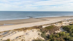 Una playa con un largo muelle que se adentra en el mar, dunas arenosas y vegetación escasa.