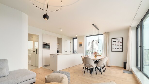 Una sala moderna con sofá, otoman y mesa redonda. Se observa un comedor con sillas y una mesa, y una cocina al fondo.