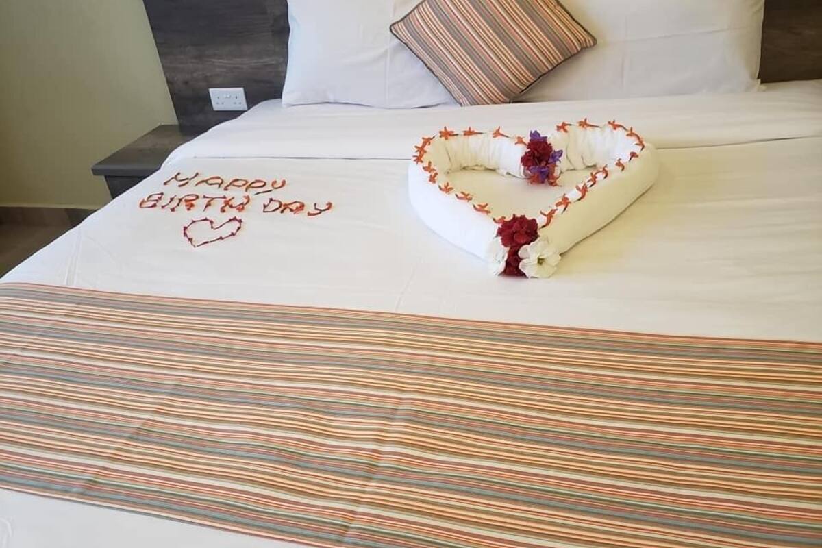 Una cama bien tendida con un arreglo floral en forma de corazón y una nota manuscrita que dice 'Feliz Cumpleaños'.
