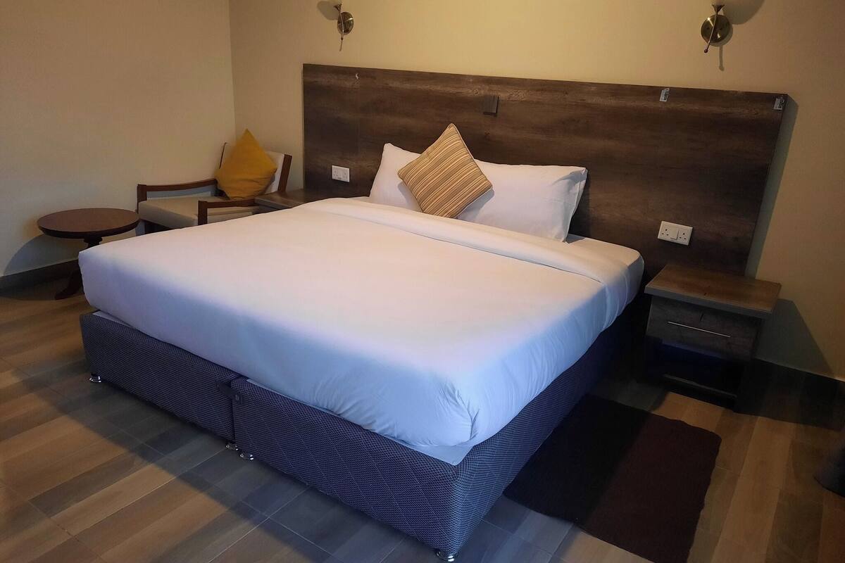 Una cama bien hecha con cabecera de madera, una mesita redonda pequeña con una silla, y una mesita de noche con una lámpara.